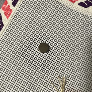 K&ouml;nnte beinhalten: Nahaufnahme einer wei&szlig;en Gobelin-Leinwand mit einem Gittermuster. Die Leinwand hat einen dekorativen Rand mit rosa und lila blattartigen Formen. Ein kleiner, runder, silberner Magnet ist auf der Leinwand zentriert. Goldfaden ist sichtbar.