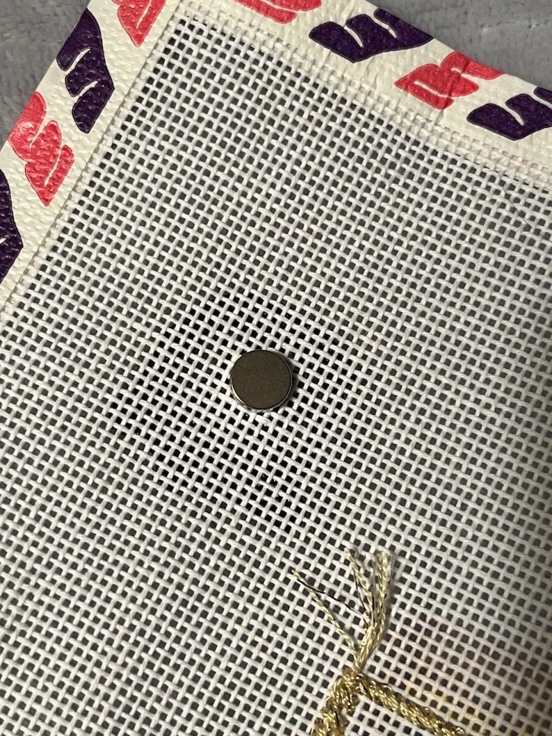 K&ouml;nnte beinhalten: Nahaufnahme einer wei&szlig;en Gobelin-Leinwand mit einem Gittermuster. Die Leinwand hat einen dekorativen Rand mit rosa und lila blattartigen Formen. Ein kleiner, runder, silberner Magnet ist auf der Leinwand zentriert. Goldfaden ist sichtbar.