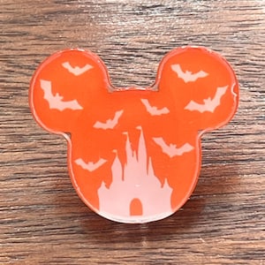 May include: Orange Mickey Mouse-shaped pin with white bat silhouettes and a castle design. The pin is a Halloween-themed accessory.