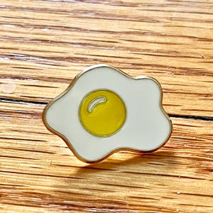 May include: A gold-edged enamel pin in the shape of a fried egg. The pin features a white egg white and a bright yellow yolk. The pin is a fun accessory for jackets, bags, or hats.