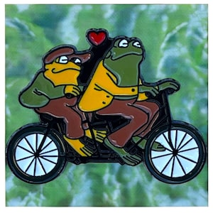 May include: Enamel pin featuring two cartoon frogs riding a tandem bicycle. The frogs are yellow and green, wearing brown and yellow outfits. A red heart floats above them. The pin has a black outline and a white background.
