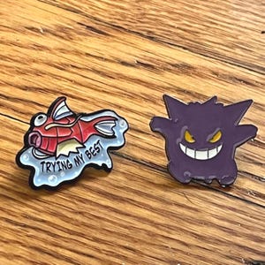 May include: Two enamel pins featuring cartoon characters. One pin depicts a red and white fish with the text "Trying My Best". The other pin is a purple ghost-like character with yellow eyes and a wide grin. Both pins are on a wooden surface.