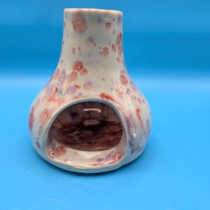 May include: A ceramic wax warmer with a wide, rounded base and a narrow, cylindrical top opening. The exterior features a white glaze with scattered, irregular spots in shades of reddish-pink and light purple, while the interior is dark.