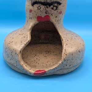 May include: A speckled beige ceramic vessel with a wide, open mouth and a hollow interior. The exterior features a hand-painted face with thick black eyebrows, small eyes, red cheeks, and a prominent red mouth positioned above the large opening.
