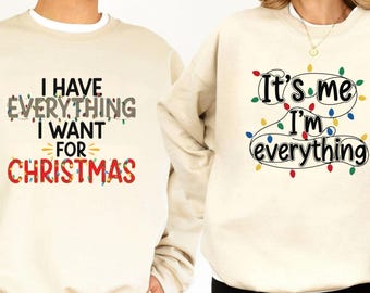 May include: Two cream-colored sweatshirts with Christmas-themed text. One says "I HAVE EVERYTHING I WANT FOR CHRISTMAS" in black, gray, and red. The other says "It's me I'm everything" with colorful Christmas lights.