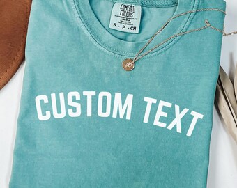 May include: A teal Comfort Colors t-shirt with the words "CUSTOM TEXT" printed in white. A gold necklace with a pendant rests on the shirt. A brown hat and dried leaves are in the background.