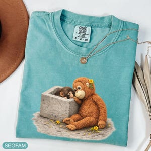 May include: A turquoise Comfort Colors t-shirt featuring a graphic of two monkeys. One monkey is sitting and the other is in a stone structure. The shirt has a small tag and a gold necklace. The shirt is size S-P-CH.
