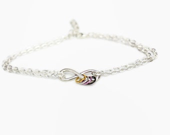 May include: A silver chain bracelet featuring an infinity symbol charm. The infinity symbol is made of silver and has a pink, purple, and gold accent.