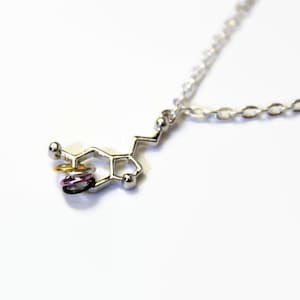 May include: Silver necklace with a silver pendant shaped like a dopamine molecule. The pendant has three small rings attached to it, one gold, one purple and one black.