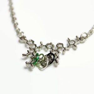 Peut inclure: Un collier en chaîne argent avec un pendentif en forme de structure chimique. Le pendentif est orné d'anneaux verts et noirs.