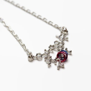 Peut inclure: Un collier en chaîne argent avec un pendentif en forme de structure moléculaire argentée. Le pendentif comporte trois petits anneaux de couleur rouge, bleu et violet.