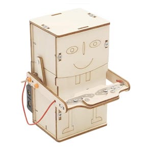 May include: A wooden robot kit with a smiling face, two square teeth, and a metal tray. The robot has a red wire and a small black box attached to its side. The robot is made of light-colored wood.