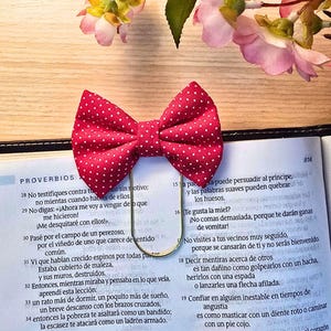 May include: Red fabric bow with white polka dots, used as a bookmark, resting on an open book. The bow is made of fabric and has a gold-colored metal clip. Pink flowers and green leaves are in the background.