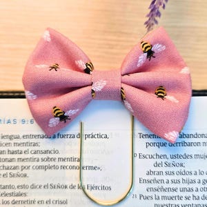 May include: A pink fabric bow with a bee pattern, attached to a gold-colored paper clip. The bow features a repeating pattern of bees with white wings and black and yellow bodies. The bow is placed on top of an open book.