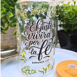 May include: A clear glass can-shaped drinking glass with a bamboo lid. The glass is decorated with green and white floral designs and the text "El justo vivirá por la fe" from Romans 1:17.