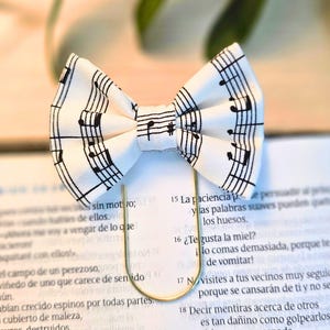 May include: A white bow tie with a musical note pattern, held by a gold-colored paper clip. The bow tie features black musical notes and rests on a book with Spanish text.