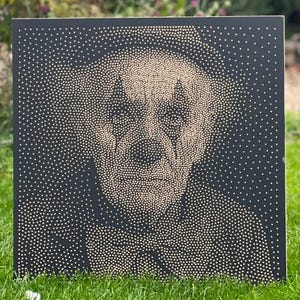 May include: Square black artwork featuring a portrait of a person's face, created with small, light-colored dots. The face has a clown-like appearance with diamond shapes around the eyes and a dark outline. The artwork is displayed on a green grassy surface.