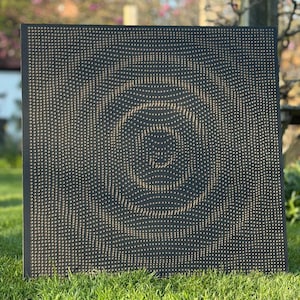 May include: A square black board covered in small, evenly spaced, gold-colored pegs. The pegs form a circular design in the center. The board is sitting on green grass.