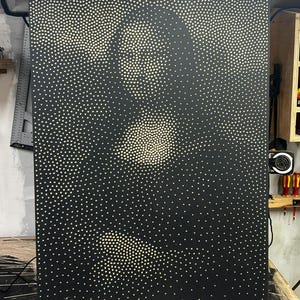 以下が含まれることがあります： 小さなオフホワイトの点がパターン化された、大きな長方形の黒いアートワーク。点は上部中央と左下に密集しており、視覚効果を生み出しています。アートワークは木製の表面に展示されています。