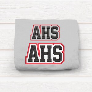 Peut inclure: Tissu gris avec deux patchs brodés noirs qui disent "AHS" dans une police de caractères audacieuse et en blocs. Les patchs ont un contour rouge.