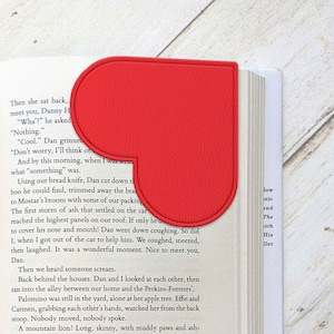 May include: Red heart-shaped bookmark with a slightly textured surface. The bookmark is placed on the corner of an open book.