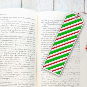 May include: A red, white, and green striped bookmark with a red tassel. The bookmark is made of fabric and has a rounded shape. The bookmark is lying on top of an open book.