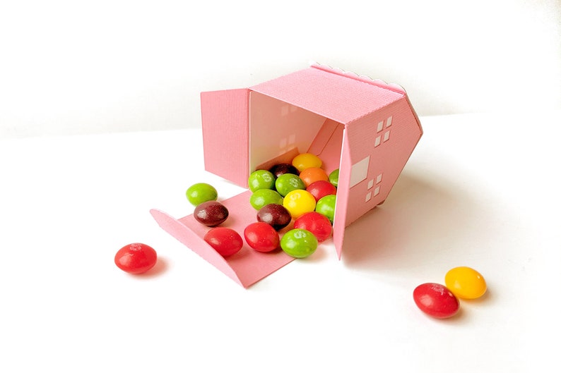 May include: A pink house-shaped box with a flap open, spilling out colorful candy. The candy is a mix of red, green, yellow, and orange.
