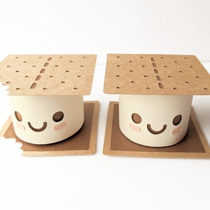 Puede incluir: Dos s'mores de cartón marrón con centros de malvavisco blancos y caras sonrientes. La galleta graham superior está ligeramente rasgada en el s'more de la izquierda.