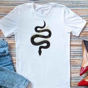 May include: White t-shirt with a black snake graphic and gold text that reads "another vaguely rumored". The shirt is paired with blue jeans and red and black high heels.