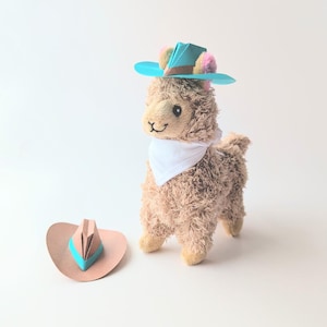 May include: A plush llama wearing a blue and brown origami cowboy hat and a white bandana. A second, matching hat lies on the surface. The llama has a smiling face and is standing upright.