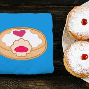 Könnte beinhalten: Ein blaues T-Shirt mit einem gestickten Donut-Motiv. Der Donut ist braun mit weißem Zuckerguss und einem rosa Herz obenauf. In der Mitte des Donuts befindet sich eine rosa Geleefüllung. Im Hintergrund befindet sich ein Teller mit Donuts mit Puderzucker und rosa Geleefüllung.