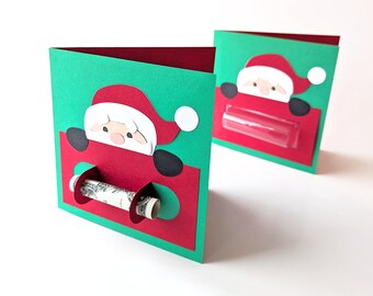 Puede incluir: Dos tarjetas de Navidad con un diseño de Papá Noel. Las tarjetas son verdes y rojas con una ventana de plástico transparente. La ventana está diseñada para contener un billete enrollado. Una tarjeta tiene un billete enrollado en la ventana.