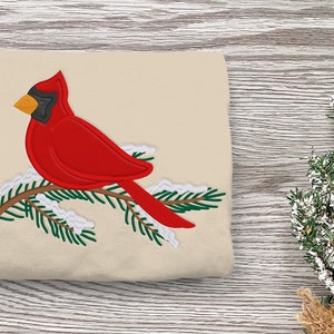 May include: A red cardinal bird perched on a green pine branch with white snow accents. The bird is facing left and has a black eye and a yellow beak.