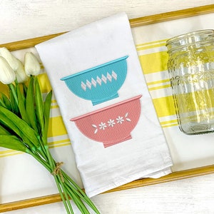 May include: A white kitchen towel with two embroidered bowls, one blue with a diamond pattern and one pink with a flower pattern. The towel is on a yellow and white striped tray with a clear glass jar and a bouquet of white tulips.