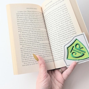 May include: An open book with text and a decorative bookmark. The bookmark is white with a green and yellow embroidered design. A hand with a gold painted fingernail holds the book open.