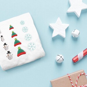 以下が含まれることがあります： 雪だるまとクリスマスハット、雪の結晶が刺繍された白いタオル。白い星、ベル、キャンディケインなど、クリスマスの飾りで囲まれており、すべて水色の背景にあります。