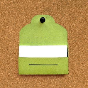 May include: A green card with a white rectangle in the center, pinned to a corkboard. The card has a rounded top and a black push pin.