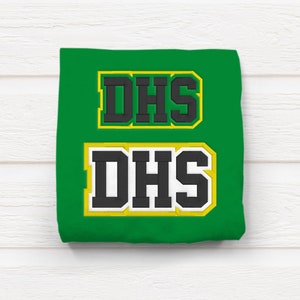May include: Green t-shirt with two black and white "DHS" patches, one above the other. The patches have a yellow outline.
