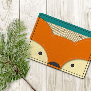 Op de afbeelding: Een kleine oranje leren portemonnee met een vosgezichtontwerp. De vos heeft zwarte ogen en een zwarte neus. De portemonnee heeft een tealblauwe rand en een beige stoffen voering.