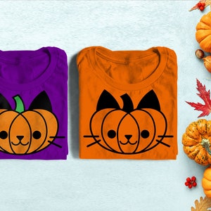 May include: Two folded t-shirts with a pumpkin-shaped cat face design. The shirt on the left is purple and the shirt on the right is orange. The design is black with green stem and white whiskers.