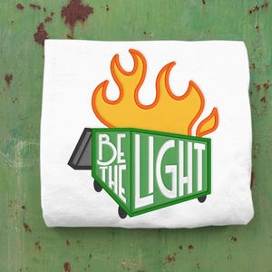 May include: A white t-shirt with a green dumpster on fire. The dumpster has the words "Be the Light" written on it.