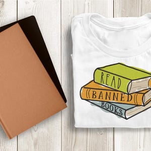 May include: A white t-shirt with a stack of three books. The top book is green and says "READ". The middle book is orange and says "(BANNED)". The bottom book is blue and says "BOOKS".
