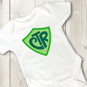 May include: White baby onesie with a green and teal shield with the letters CTR inside.