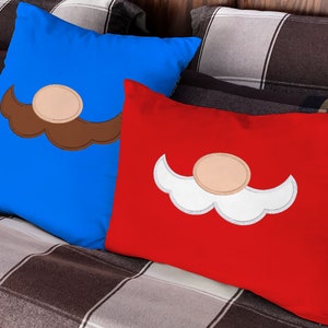 Op de afbeelding: Twee decoratieve kussens met geborduurde snorren. Een kussen is blauw met een bruine snor en de andere is rood met een witte snor.