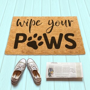 Puede incluir: Una alfombra de coco marrón con el texto "wipe your paws" en letras negras. El texto incluye un gráfico de huella de pata.
