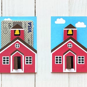 Puede incluir: Dos porta tarjetas con forma de escuela roja con fondo de cielo azul. El porta tarjetas de la izquierda tiene una tarjeta de regalo insertada. El porta tarjetas de la derecha está vacío.