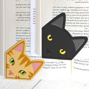 May include: Two cat-shaped bookmarks rest on open book pages. One is black with yellow eyes, the other is orange with green eyes. The bookmarks are made of a soft material.