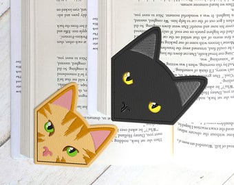 May include: Two cat-shaped bookmarks rest on open book pages. One is black with yellow eyes, the other is orange with green eyes. The bookmarks are made of a soft material.