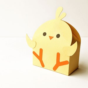 Puede incluir: Una caja de papel de pollito amarillo con patas naranjas y un pico. El pollito tiene ojos negros y una expresión linda.