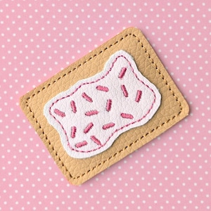 May include: A tan leather patch in the shape of a rectangle with a stitched border. A white patch with pink sprinkles is stitched on top. The background is pink with white polka dots.